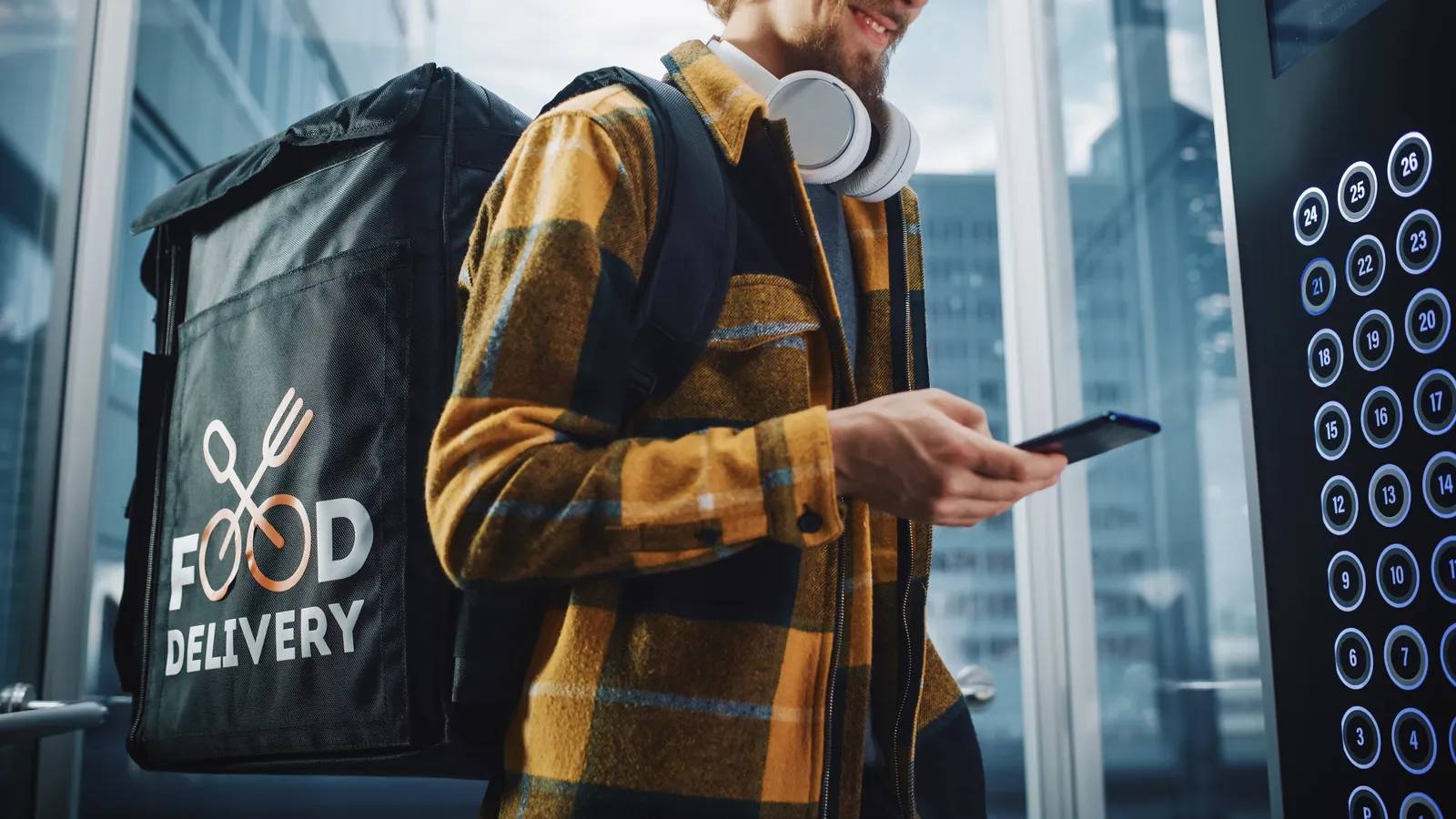 A smiling food delivery worker wearing a yellow plaid shirt, headphones, and a large backpack enters an elevator while checking a smartphone. The backpack has a “Food Delivery” logo on it. Elevator buttons are visible on the right.