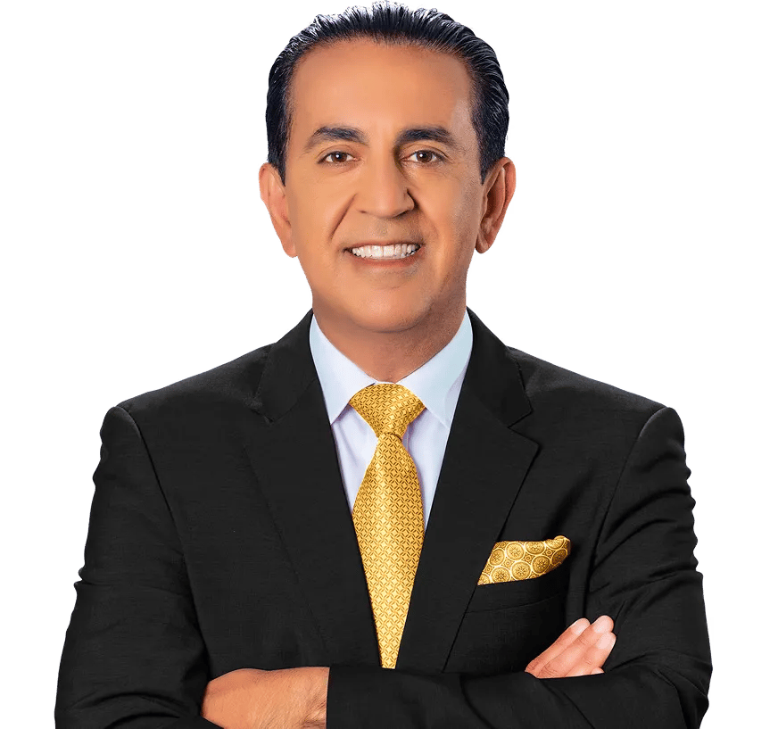 A man in a black suit, white shirt, and yellow patterned tie stands with arms crossed, smiling at the camera against a light background. He has dark hair slicked back and is wearing a matching yellow pocket square.