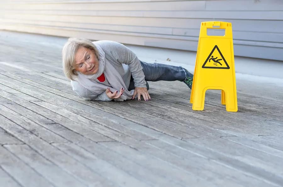 An older woman lies on a wooden floor after a fall, looking distressed. A yellow caution sign with a slipping person icon is nearby, highlighting the risk of personal injury from slippery surfaces.