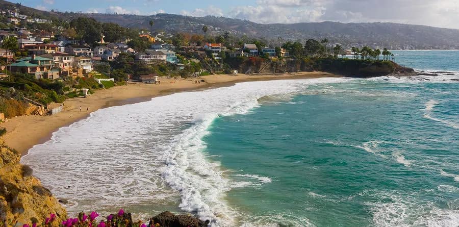 A scenic coastal view of a sandy beach bordered by turquoise waves and rocky cliffs, with houses and greenery on the hillsides under a partly cloudy sky—perfect for relaxing or meeting with personal injury attorneys in a serene setting.