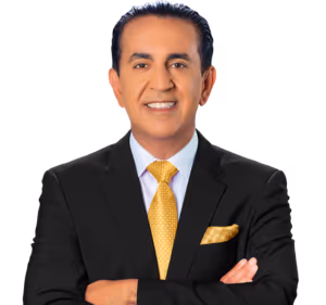 A man in a black suit, white shirt, and yellow patterned tie stands with arms crossed, smiling at the camera against a light background. He has dark hair slicked back and is wearing a matching yellow pocket square.