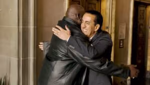 Two men, one in a black leather jacket and the other in a dark suit, are smiling and embracing warmly in an ornate indoor setting—perhaps celebrating a big win as personal injury lawyers.