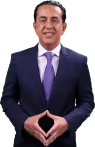 A man wearing a dark blue suit, white shirt, and purple patterned tie stands smiling with his hands together in front of him, fingers touching to form a triangle.