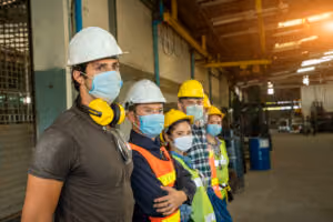A group of factory workers wearing hard hats, safety vests, and face masks stand side by side inside an industrial warehouse, looking in the same direction—much like a team of personal injury attorneys ready to advocate for workplace safety.