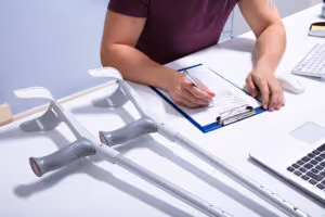 A person sitting at a desk fills out a form on a clipboard, with crutches resting on the table nearby—suggesting they may be dealing with a personal injury—next to a laptop and keyboard.