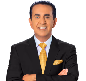 A man in a black suit, white shirt, and yellow patterned tie stands with arms crossed, smiling at the camera against a light background. He has dark hair slicked back and is wearing a matching yellow pocket square.